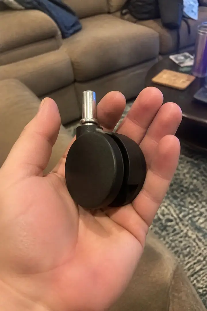 A black twin-wheel office chair caster held in an open palm in front of a sofa